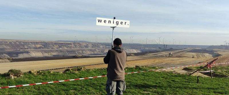 Mann mit Schild "Weniger." - Im Hintergrund Kohleabbau